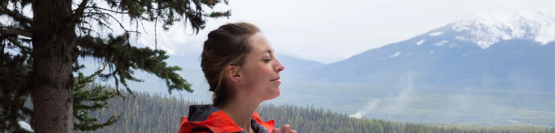 woman in red jacket sits in wooded area with body of water in background