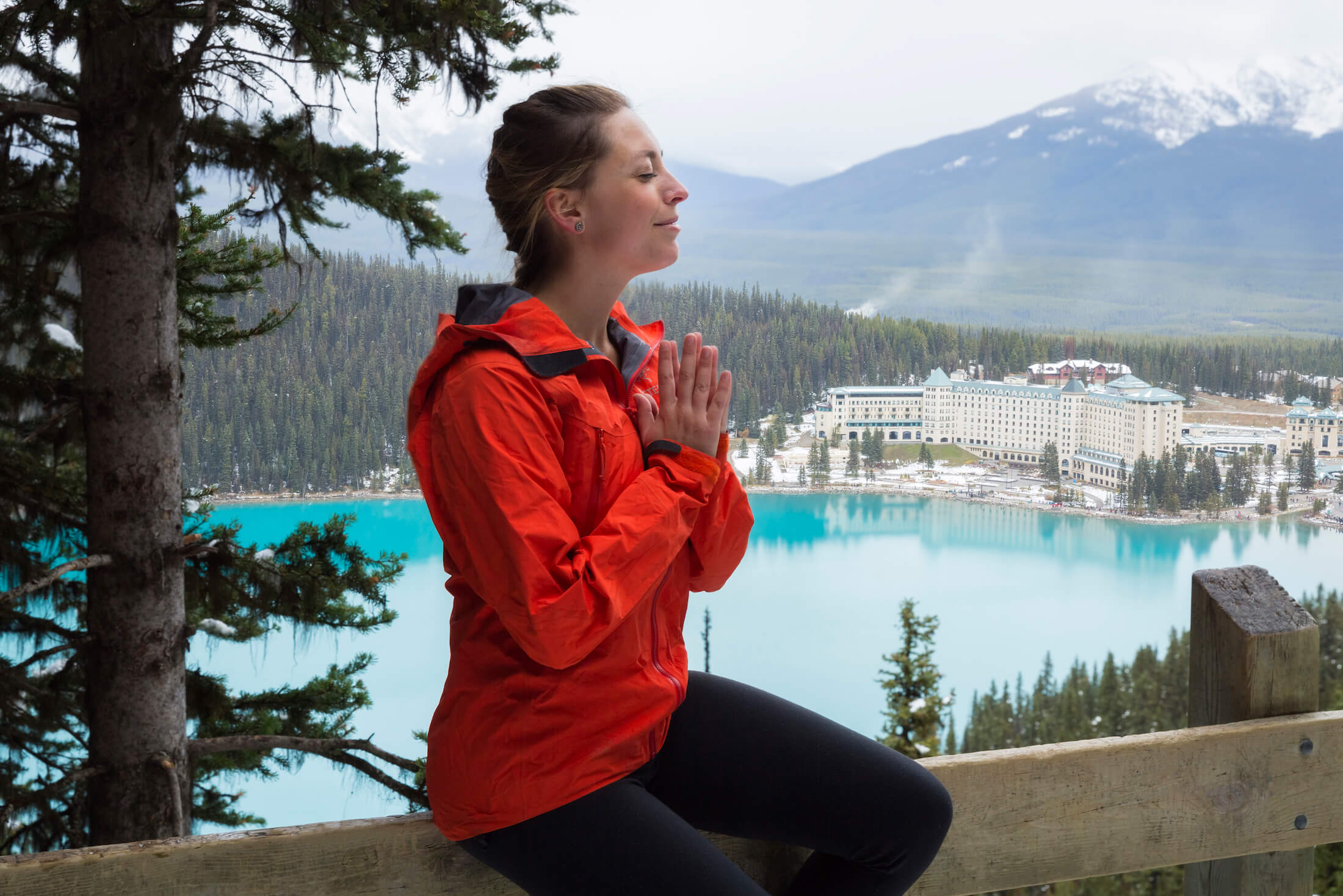 woman in red jacket sits in wooded area with body of water in background