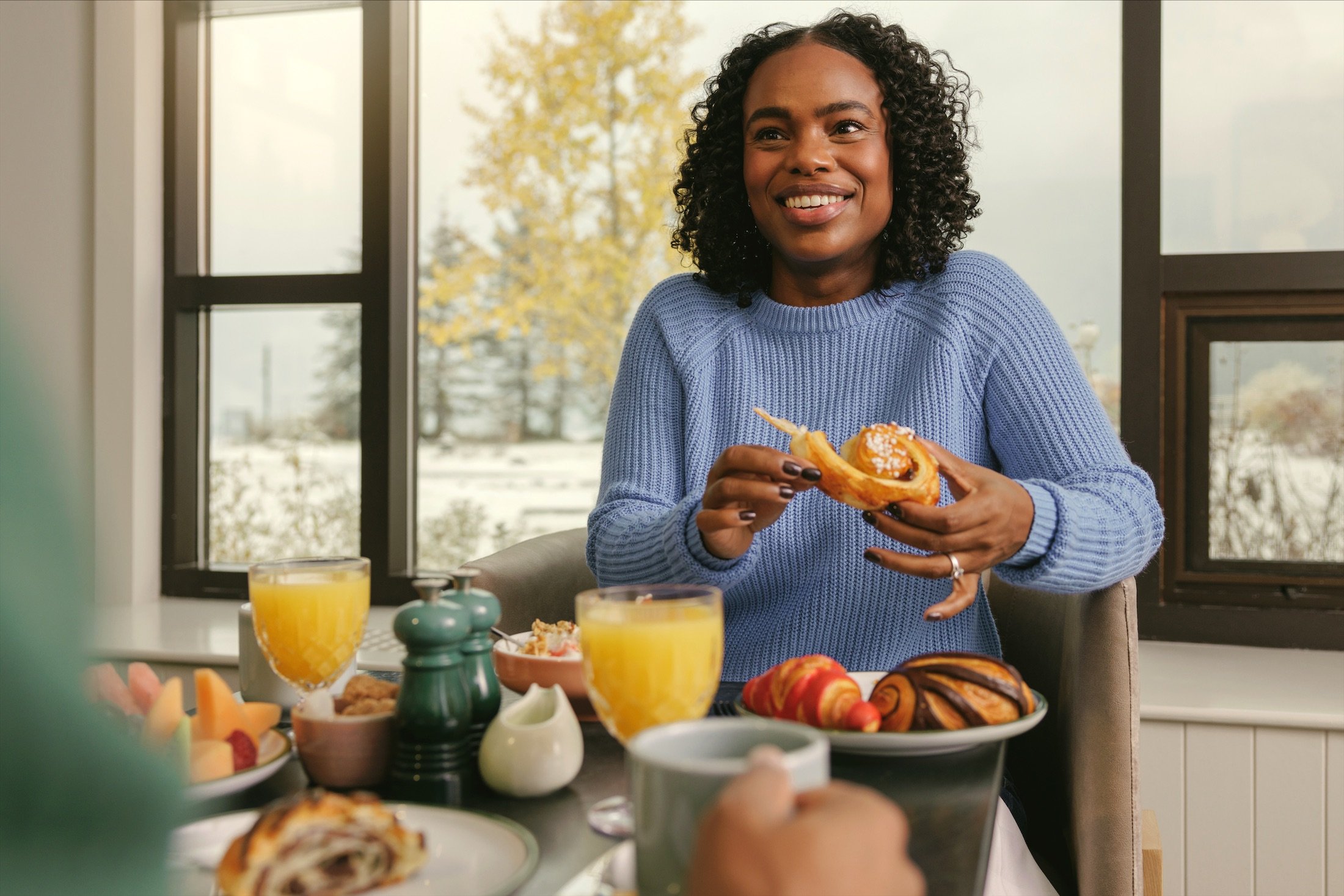 A woman at a breakfast table loaded with fruit, juice, and pastries