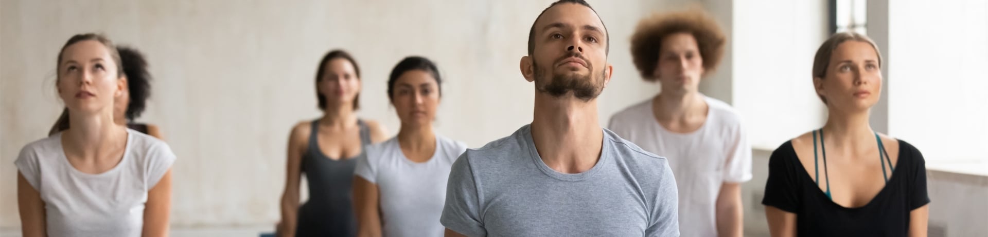 Diverse people wearing sportswear doing cobra exercise at group lesson, practicing yoga on mats, stretching in Bhujangasana pose, working out in modern yoga studio with young male instructor