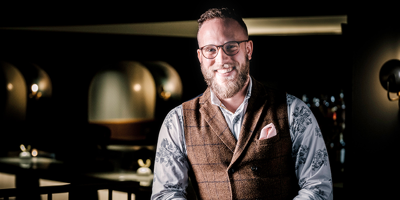 a man wearing glasses, vest, and light coloured shirt smiles