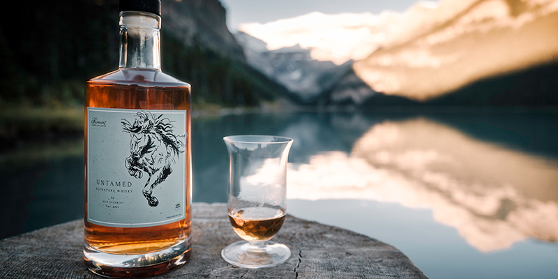 bottle of whiskey beside a tasting glass sit in front of a body of water