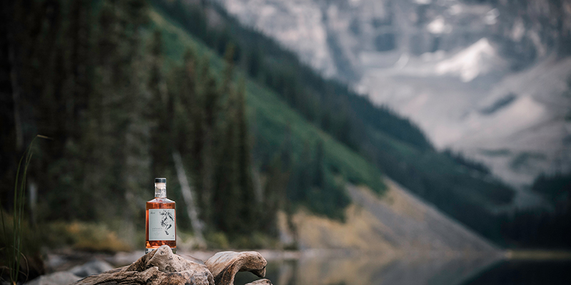 bottle of whiskey beside a tasting glass sit in front of a moutain range