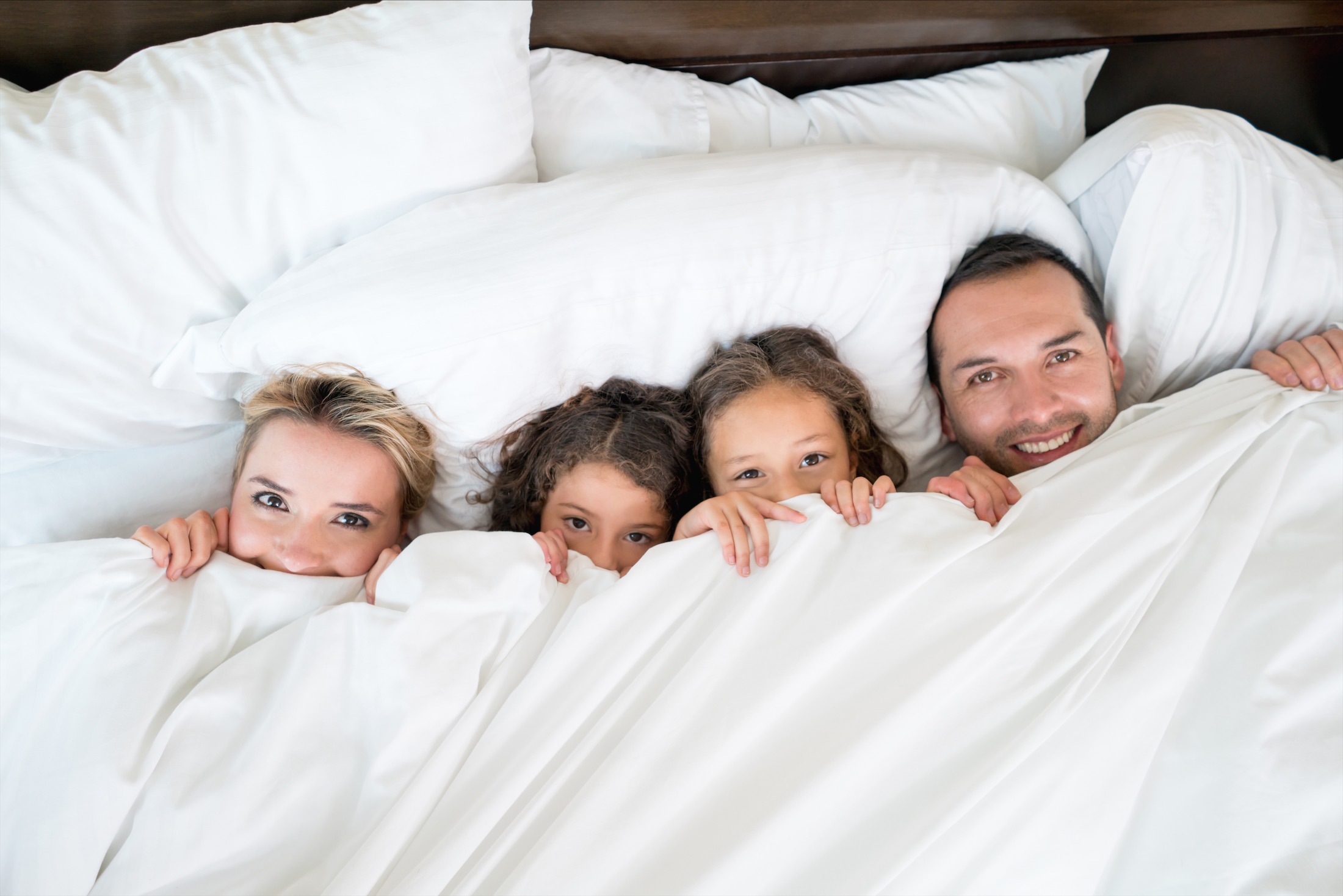 family of four lay on bed with white sheets pulled up to their chins