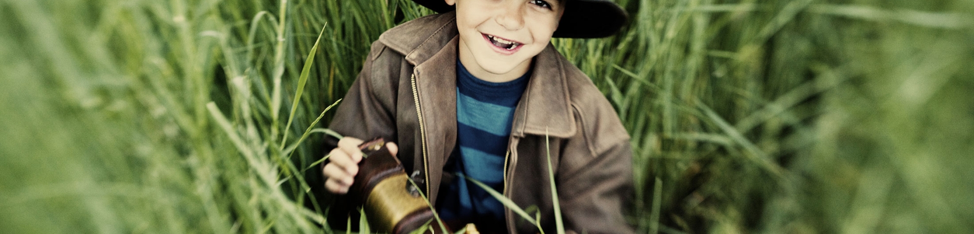 boy sitting in long grass with a small treasure chest in a dark coloured brimmed hat